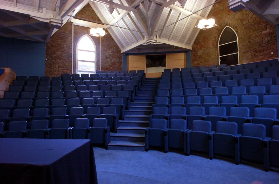 Tiered auditorium  with wooden vaulted ceiling and arched windows and blue colour theme.