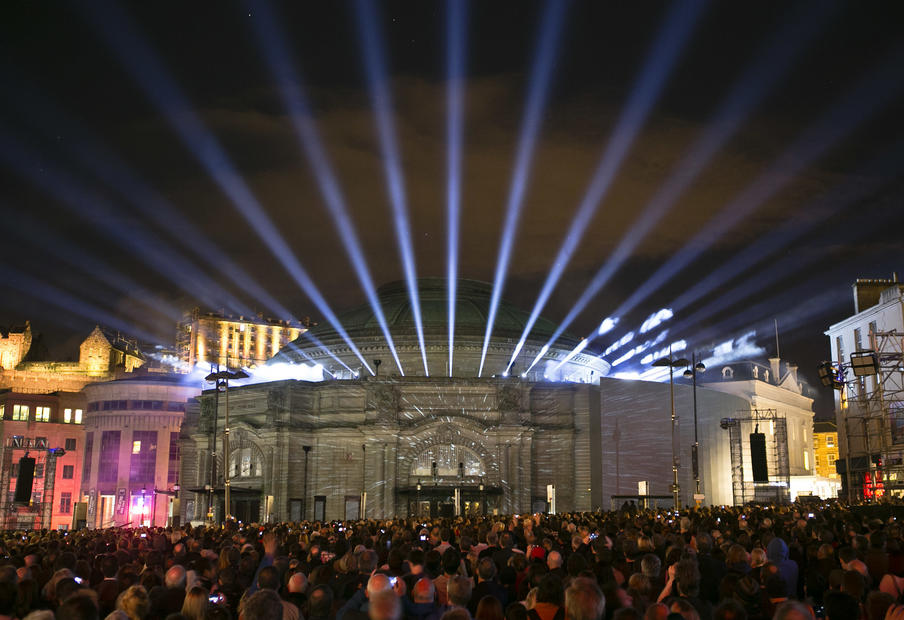 Multiple spotlights shine from the top of the domed roof of Edinburgh's Usher Hall during a large scale public projection event. A sea of people are in front of the building.