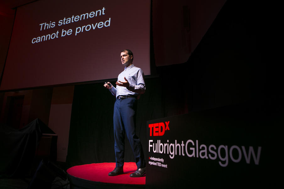 A white man wearing blue shirt and trousers presents on a dark spotlit stage at a TED talk event.
