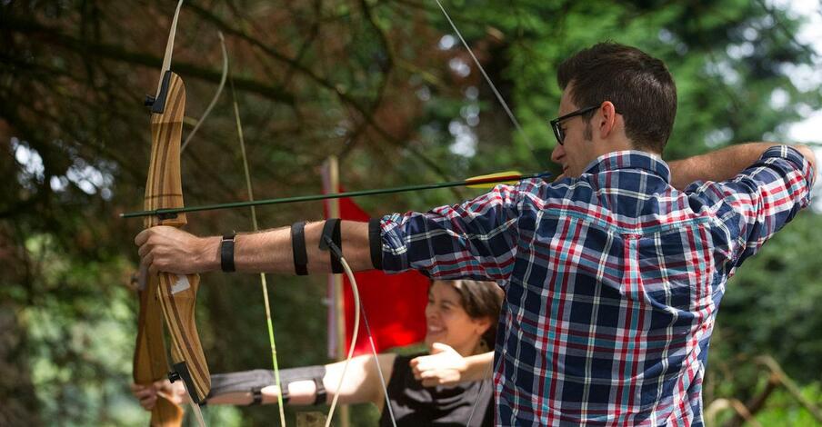 A man and a woman practice archery outdoors, aiming their recurve bows at a target. The man wears a plaid shirt, and the woman smiles in the background. Trees are visible around them.