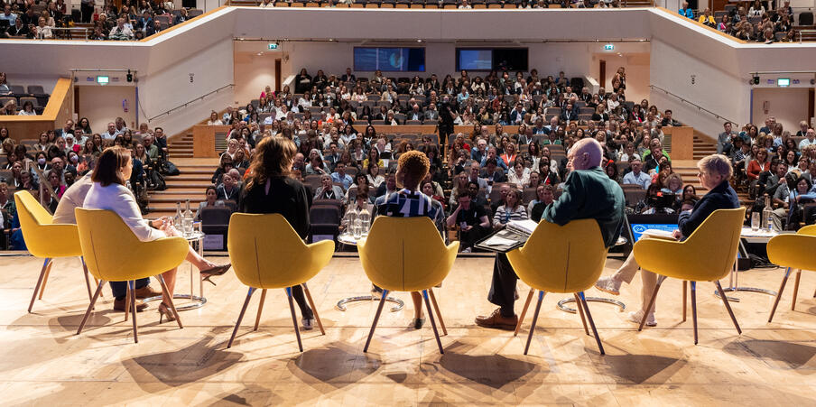 Image is a view from behind 5 speakers on stage in yellow chairs, looking out into a full crowd at a lecture.