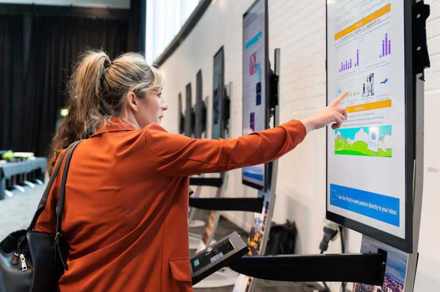 Woman in an orange jacket with blonde hair pointing at data tables on a digital screen.
