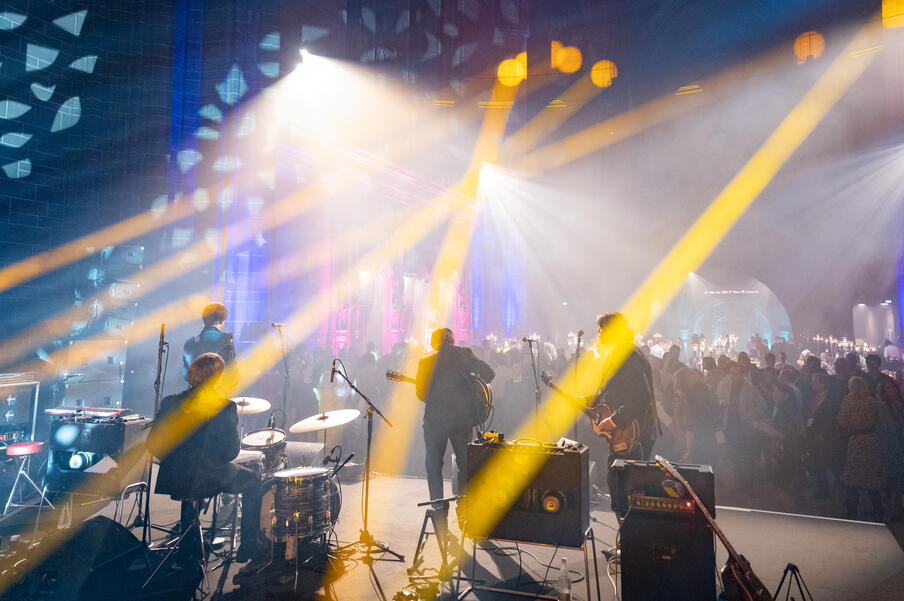 View from back of stage. Image shows a band of four men performing on stage with drums an guitars, looking out onto a busy crowd with yellow and blue strobe lights across the picture.