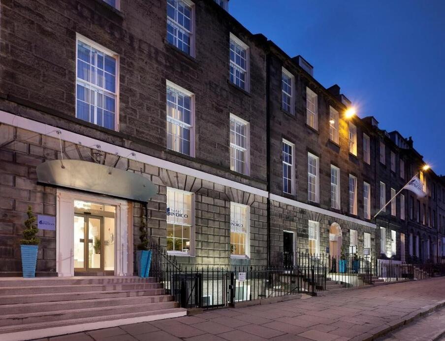 Nighttime picture of the front of the hotel. the hotel front is grey stone with windows that have Hotel Indigo branding on them.