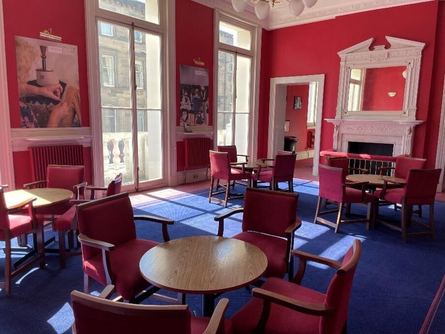 elegant bar room with multiple four seat tables on a blue carpet. The room has floor to ceiling windows, red walls and a mirror fireplace.