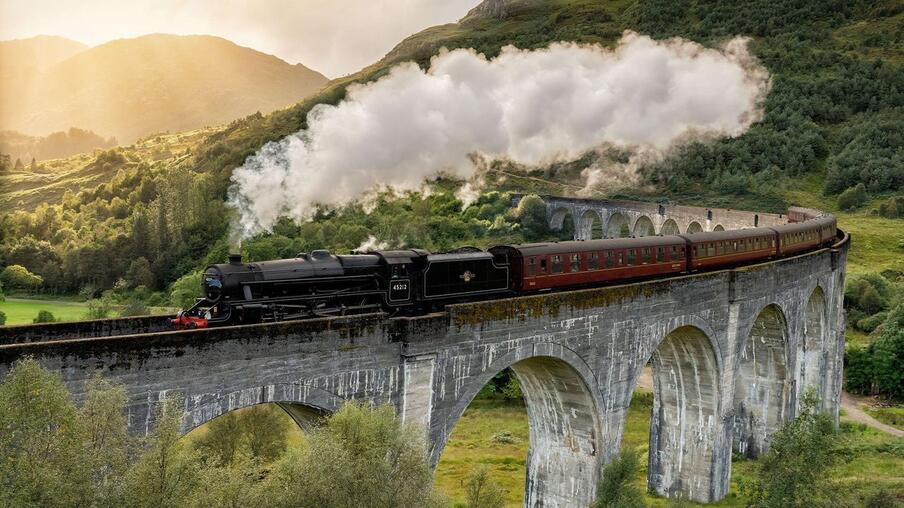 Black steam train crossing the Glenfinnan Viaduct, with rolling hills in the backround.