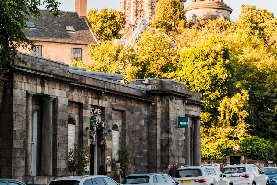 Stone building with arched windows in warm sunlight, surrounded by lush greenery. Cars parked in front, creating a tranquil, inviting atmosphere.