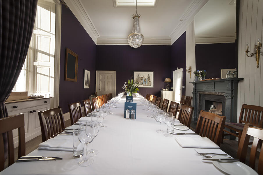 Elegant dining room with a long table set for a formal meal, featuring white tablecloth, wine glasses, and silverware. Purple walls and a chandelier add sophistication.
