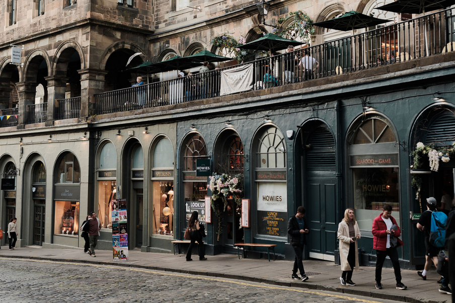 Historic cobblestone street with arched storefronts and a lively balcony café above. Stylish pedestrians and a relaxed, charming atmosphere.