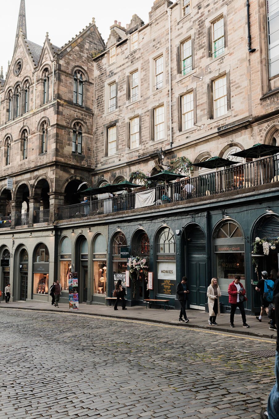 Historic stone building with arched windows and a quaint balcony above shopfronts. People stroll on cobblestone street, creating a lively atmosphere.
