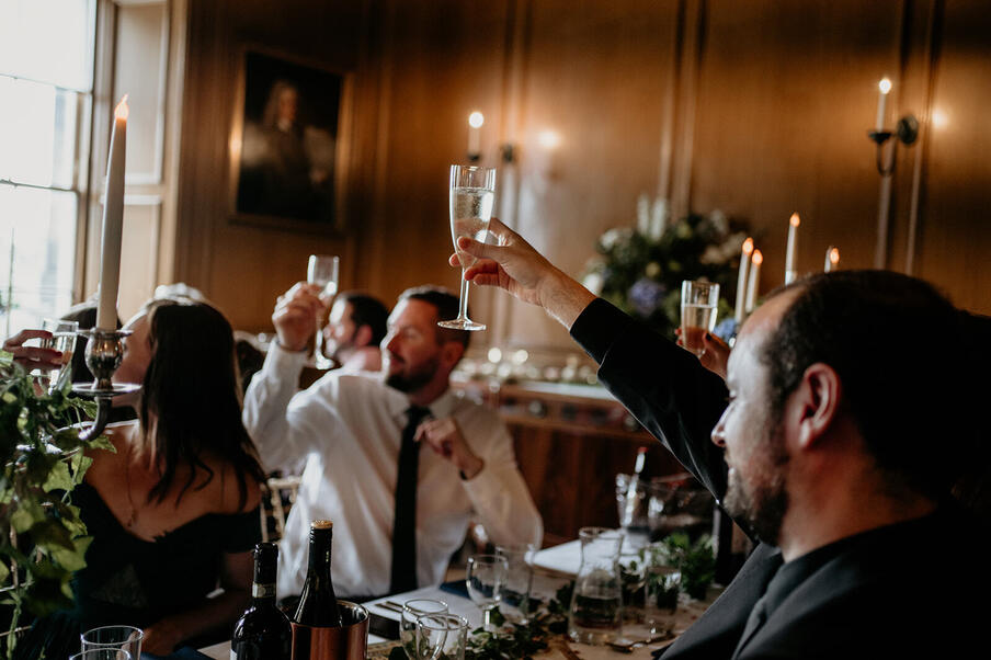 People raise champagne glasses in a toast at an elegant dinner with dim lighting, candles, and wood-panelled walls, creating a celebratory atmosphere.