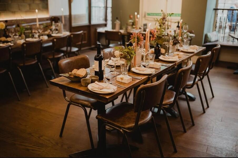 Elegant dining room set for a formal gathering. A long wooden table with neatly arranged plates, glasses, cutlery, wine bottles, candles, and flowers. Warm lighting adds a cosy atmosphere.