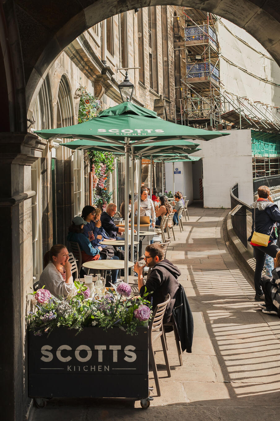 Outdoor seating at a café with green umbrellas and a sign reading "Scotts Kitchen." Patrons relax and chat on a sunny day, casting shadows.