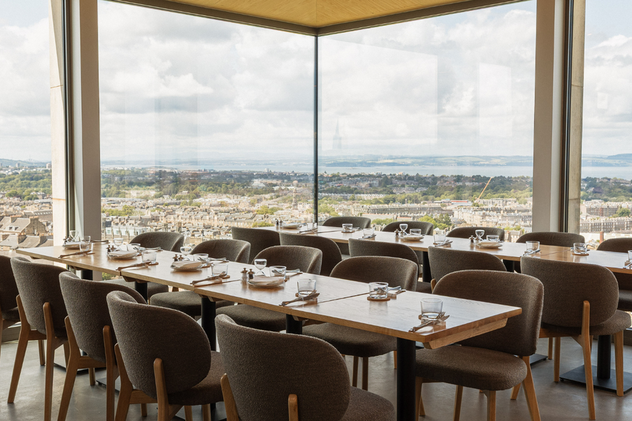 Modern dining room with large windows offering a panoramic cityscape view. Neatly arranged tables and cushioned chairs create an inviting atmosphere.