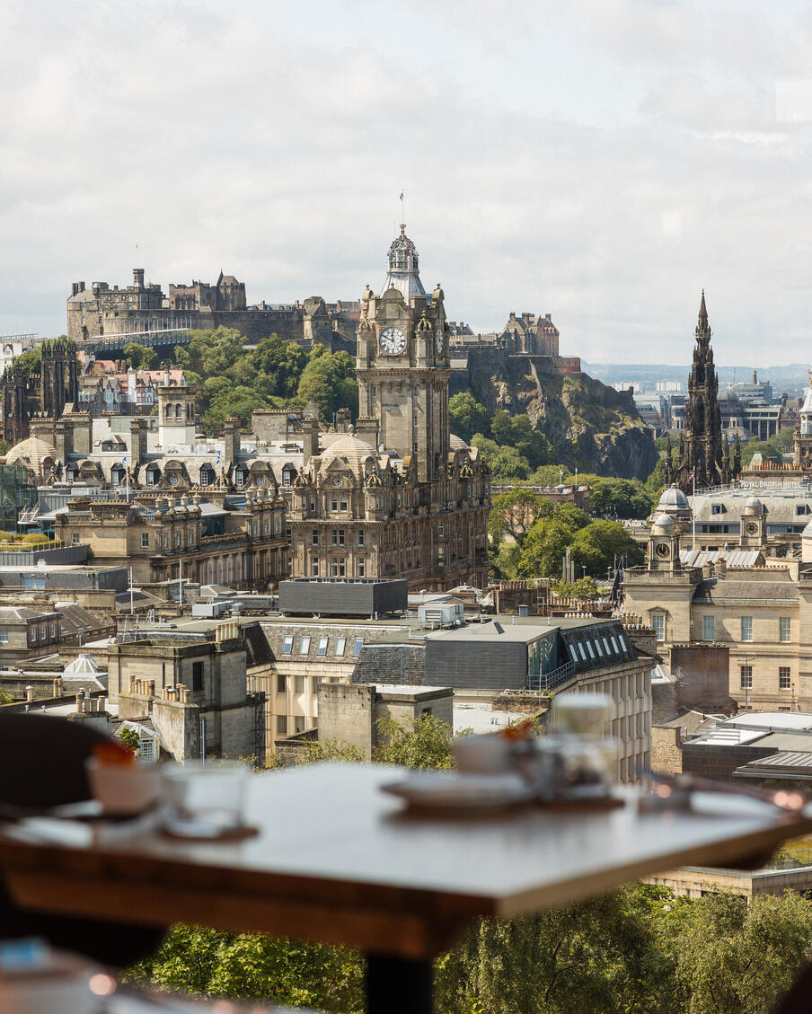 View of a cityscape dominated by an ornate clock tower, historic buildings, and lush greenery in the background. Foreground shows a blurred dining table.