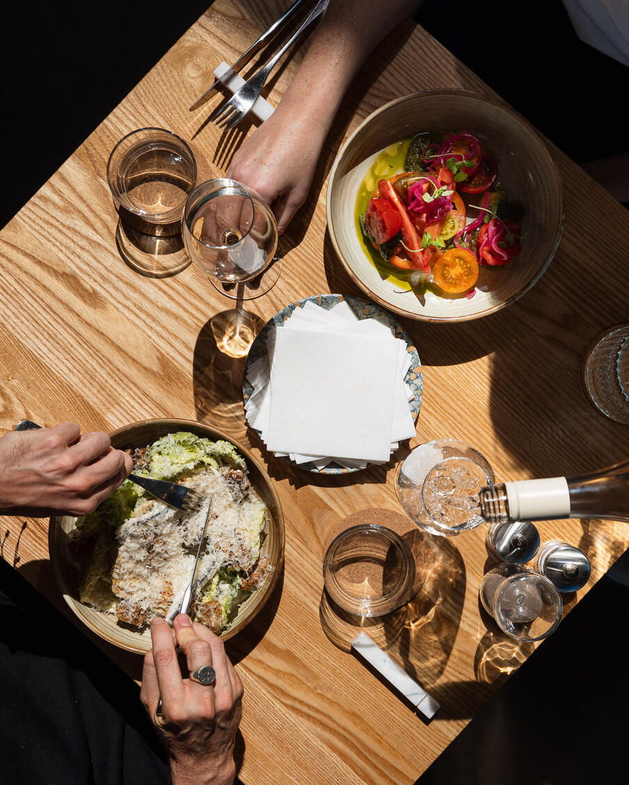 Two people share a meal at a sunlit wooden table. One person slices a dish topped with grated cheese, while the other reaches for a colorful salad. Glasses of wine and water reflect the light, creating a warm and inviting atmosphere.