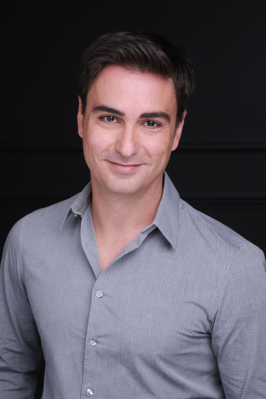 Image of a man with dark hair wearing a grey shirt in front of a black background.