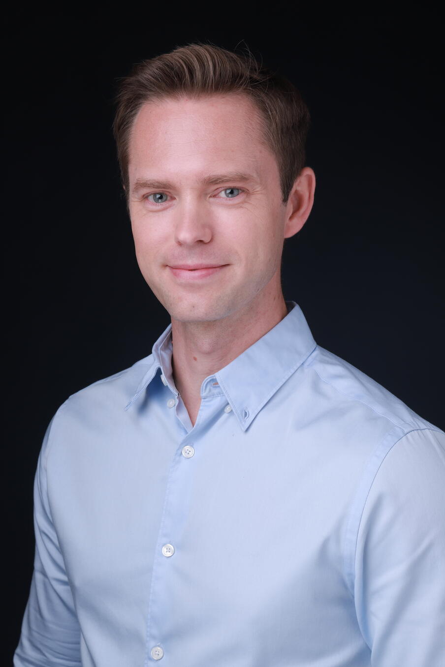 Image of a man with brown hair wearing a blue shirt in front of a black background.