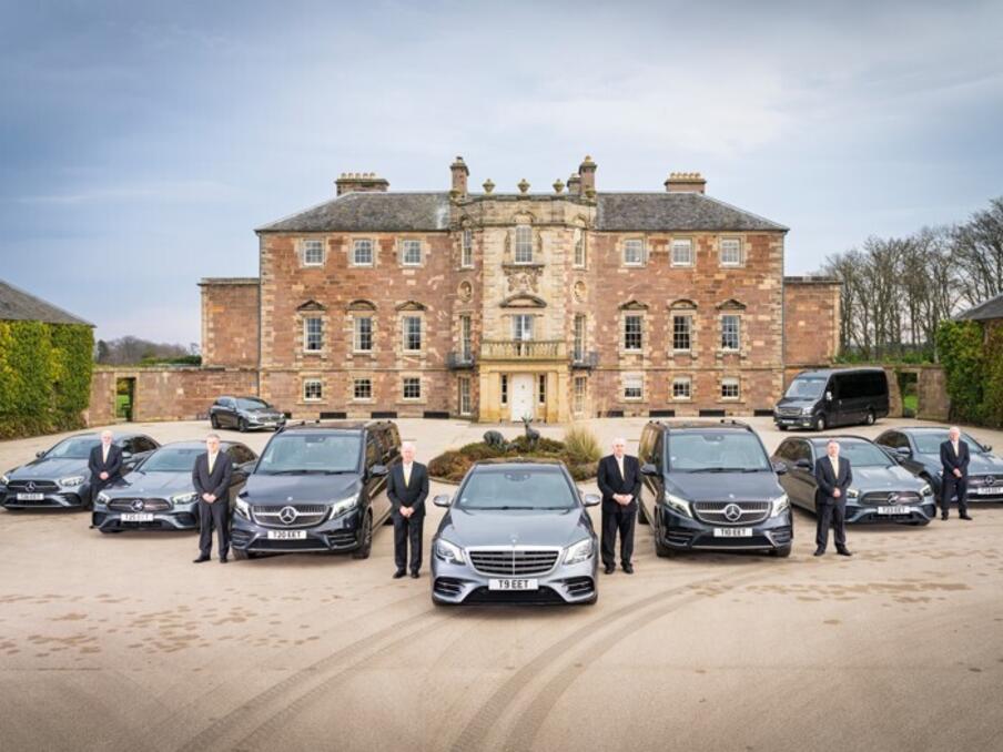9 black cars and mini buses outside of a manor house with staff in suits beside the vehicles